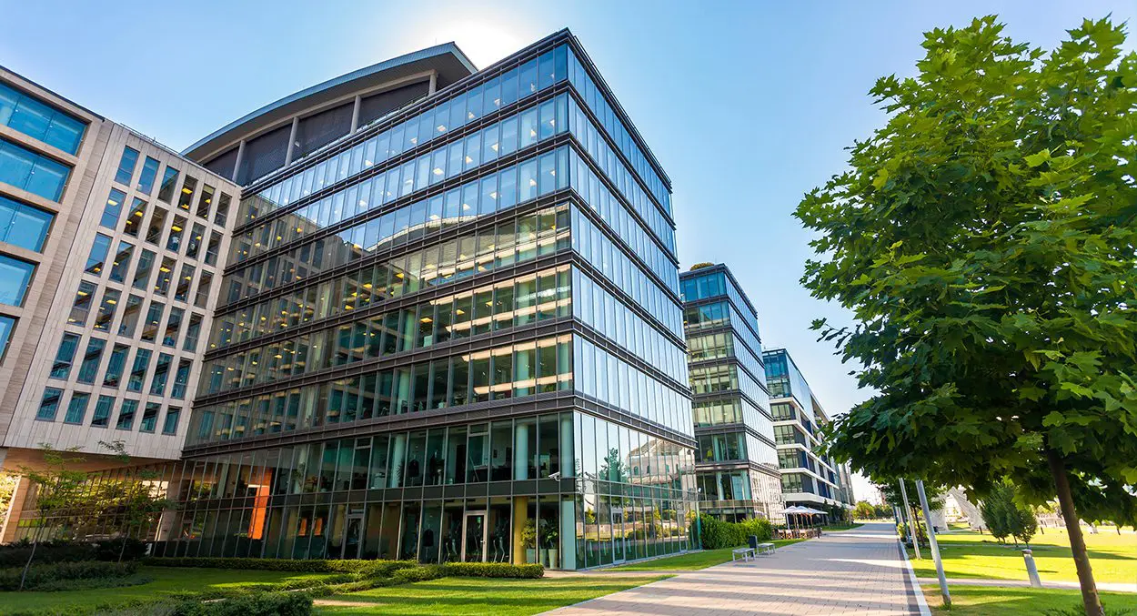 Modern glass office buildings with multiple stories, surrounded by green lawns and trees, under a clear blue sky.