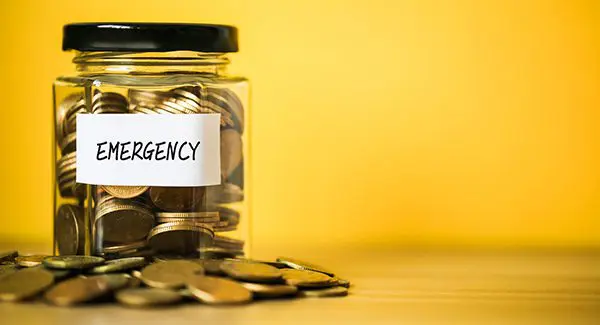 A glass jar labeled "EMERGENCY" is filled with coins and surrounded by more coins on a wooden surface, with a yellow background.