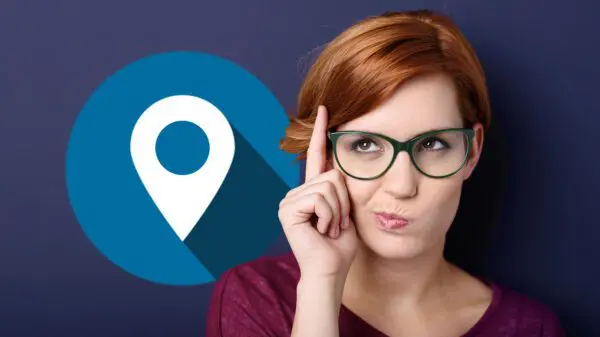 Woman with glasses touches her forehead thoughtfully in front of a blue map location pin icon on a dark background.