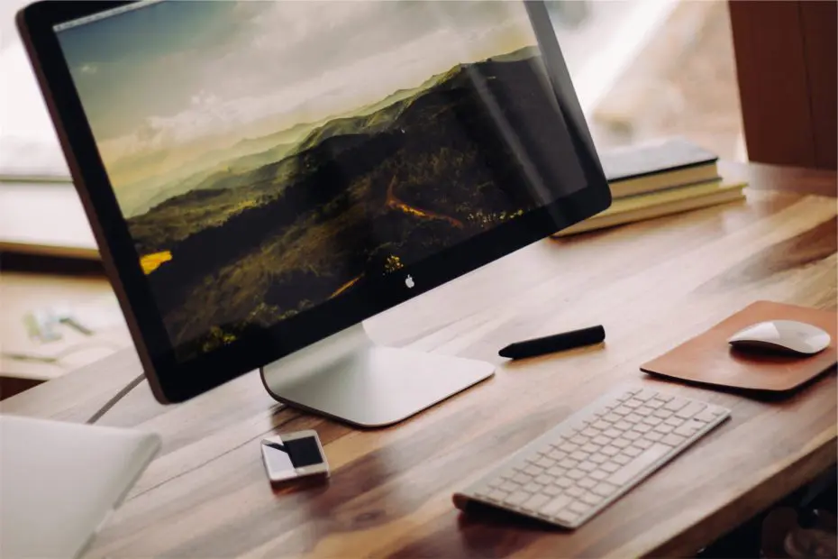 A desktop computer with a landscape on the screen sits on a wooden desk, alongside a keyboard, mouse, stylus, USB drive, and stacked notebooks.