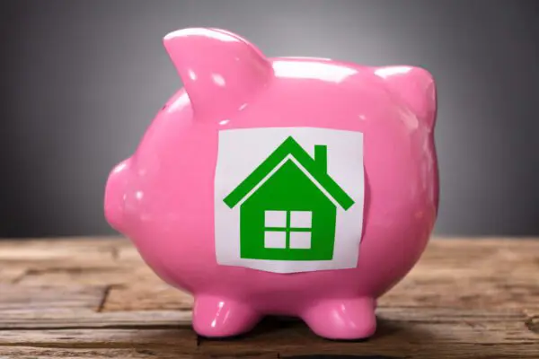 A pink piggy bank with a green house symbol on its side sits on a wooden surface against a gray background.