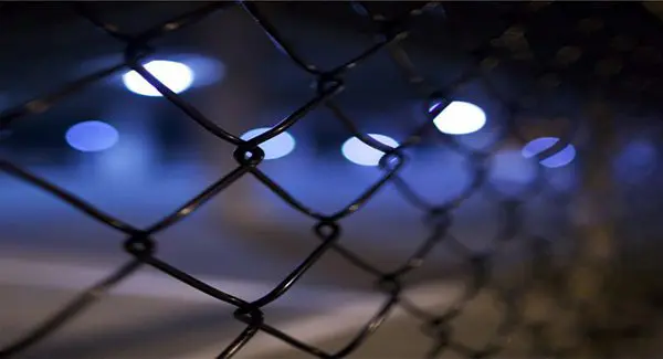 Close-up of a chain-link fence with blurred blue and white lights in the background.