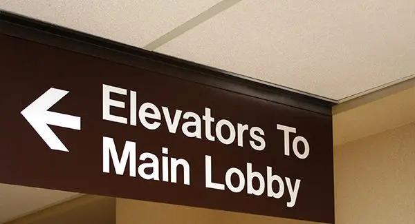 Brown sign with white text and a left arrow reads "Elevators To Main Lobby," hanging from a ceiling in an indoor setting.