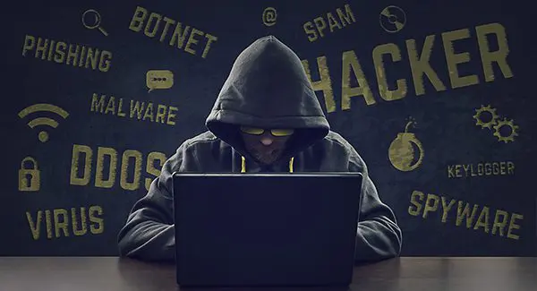A person in a hooded sweatshirt using a laptop, with cybersecurity threat words like "Hacker," "Spam," "Virus," and "Phishing" in the background.