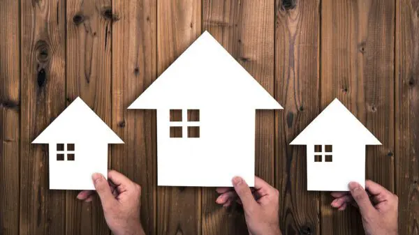 Three hands hold three white paper cutouts of houses of different sizes against a wooden background.