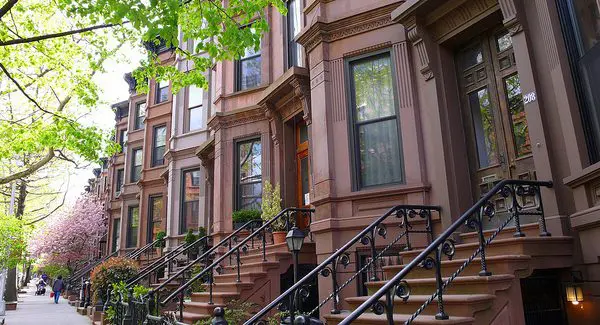 A row of brownstone townhouses with stoops and railings lines a leafy residential street on a sunny day.