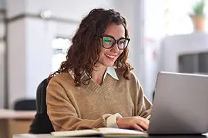 A woman with curly hair and glasses is smiling while working on a laptop at a desk in a bright office setting.