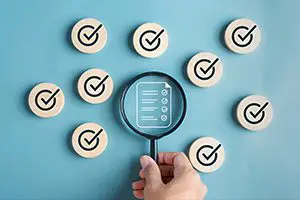 A hand holds a magnifying glass over a checklist icon, surrounded by wooden circles with check marks on a blue background.