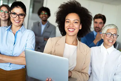 A diverse group of six professionals, including women and men, stand together in an office setting, smiling confidently. One woman holds a laptop.