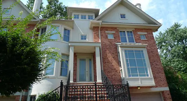 Brick and stucco two-story house with large front windows, white trim, and a black wrought iron railing along the front steps, surrounded by trees.