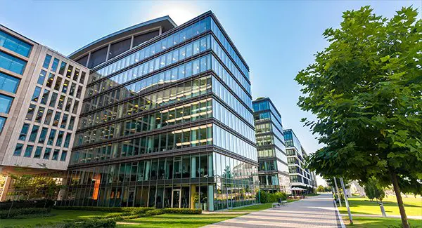 Modern glass office buildings with multiple floors, surrounded by green lawns and trees, under a clear blue sky.