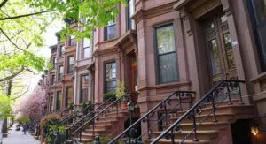 Row of brownstone townhouses with iron railings and steps, trees with green leaves, and a sidewalk with pedestrians on a sunny day.