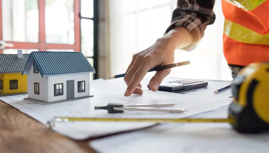 A person in a plaid shirt and safety vest reviews architectural plans on a desk, checking for potential construction defects, with model houses, a calculator, and measuring tools visible.