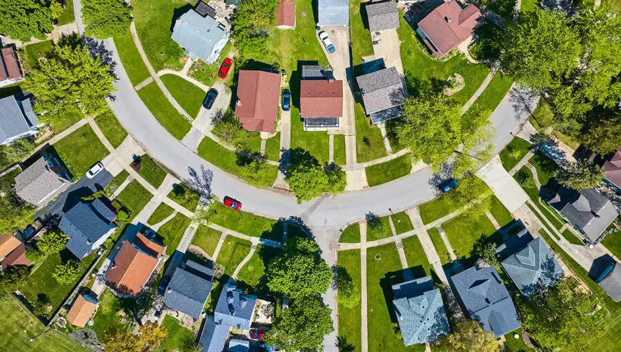 Aerial view of a suburban neighborhood showing curved streets, houses with yards, driveways, and parked cars.