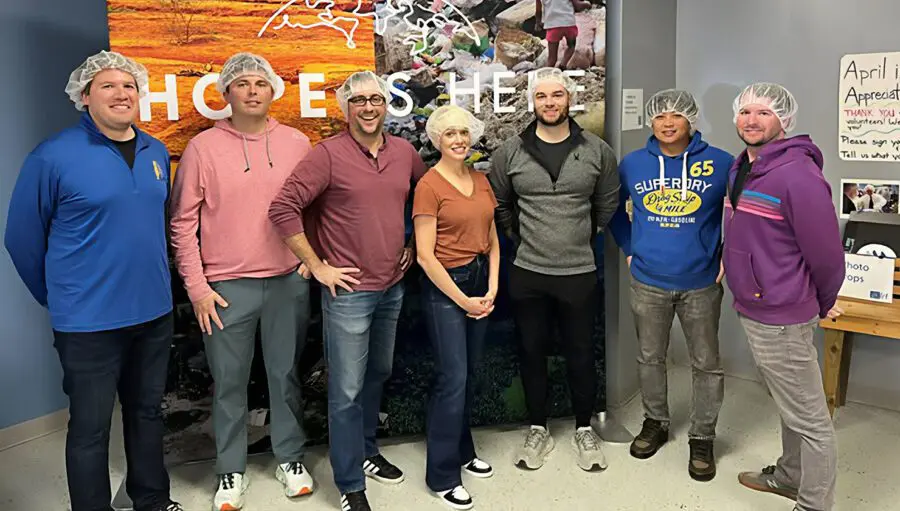 Seven people wearing hair nets stand indoors in front of a “Hope is Here” sign, posing for a group photo.