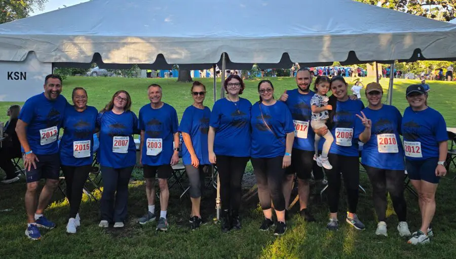 A group of eleven people, all wearing blue shirts and numbered race bibs, pose together in front of a tent at an outdoor event. One person is holding a child.