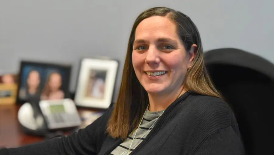 A woman with straight brown hair and a black sweater sits at a desk, smiling at the camera. Blurred family photos and an office phone are visible in the background.