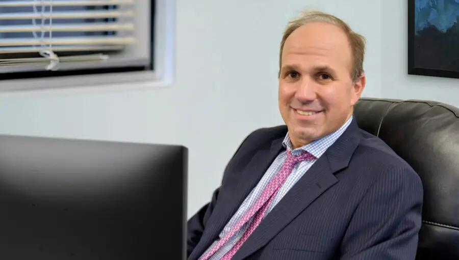 A man in a suit and tie sits at an office desk, smiling at the camera. A window with blinds and part of a framed picture are visible in the background.