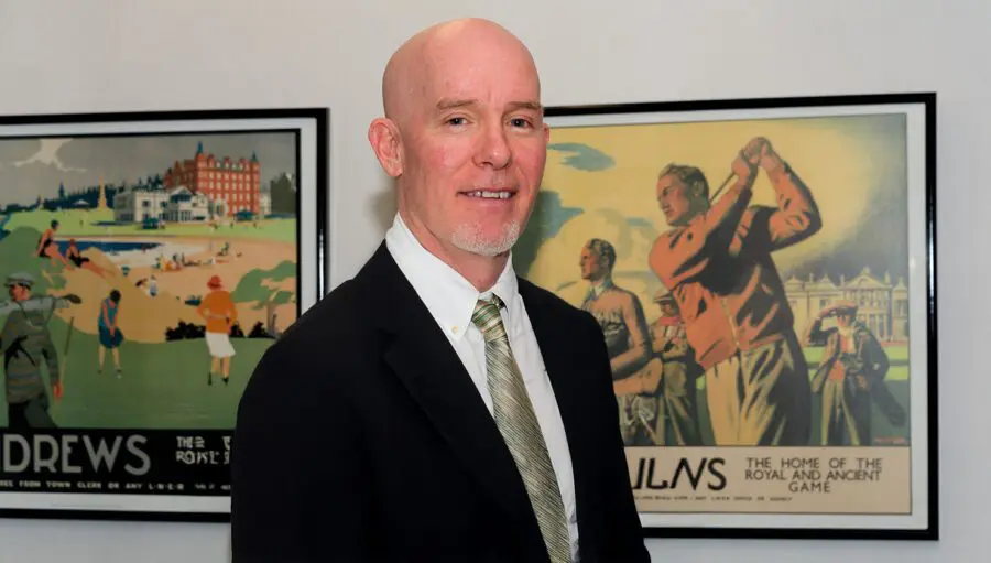 A bald man in a suit and tie stands in front of two vintage golf-themed posters on a wall.