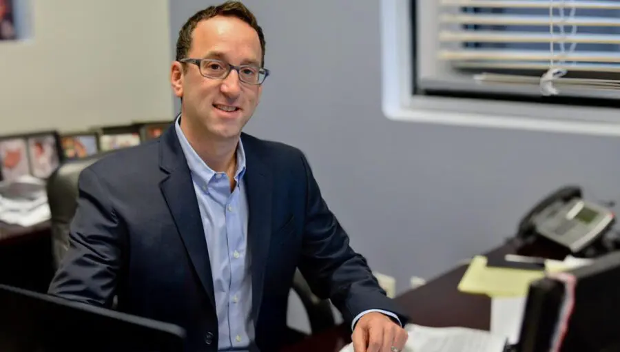 A man in a suit sits at a desk in an office, smiling at the camera. Papers, a computer, and a phone are on the desk, with family photos in the background.