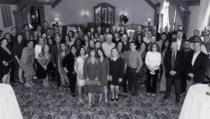 A large group of professionally dressed people pose together for a group photo in a well-lit room with ornate decor and carpet.