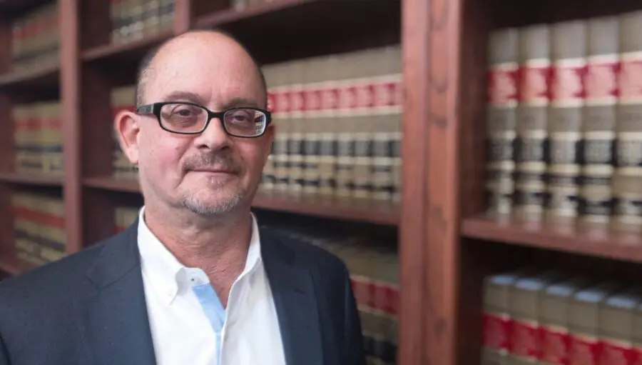 A man wearing glasses and a suit stands in front of shelves filled with legal books.
