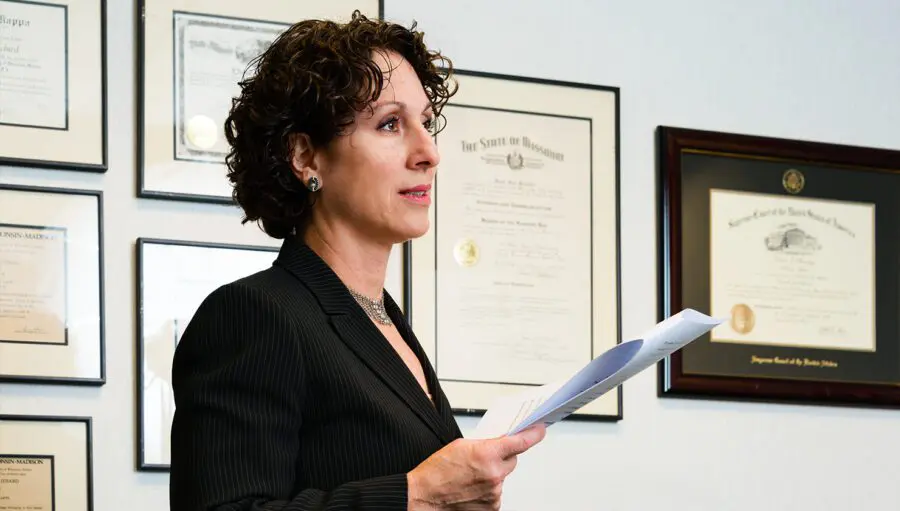 A woman in a suit holds papers while standing in front of a wall with framed certificates and diplomas.