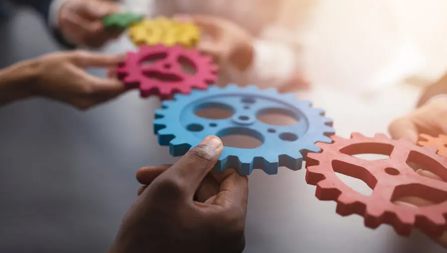 Several hands hold colorful interlocking gears, symbolizing teamwork and collaboration in a well-lit indoor setting.