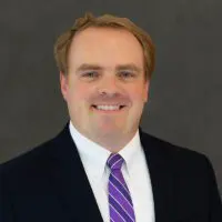 A man in a dark suit, white shirt, and purple striped tie smiles at the camera against a plain dark background.