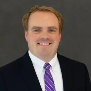 A man in a dark suit, white shirt, and purple striped tie smiles at the camera against a plain dark background.