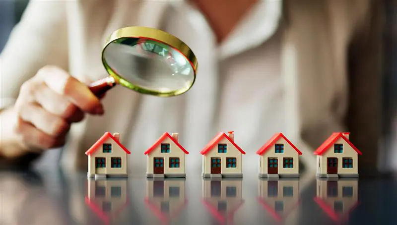 A person holds a magnifying glass over a row of five small model houses on a reflective surface, examining them closely.