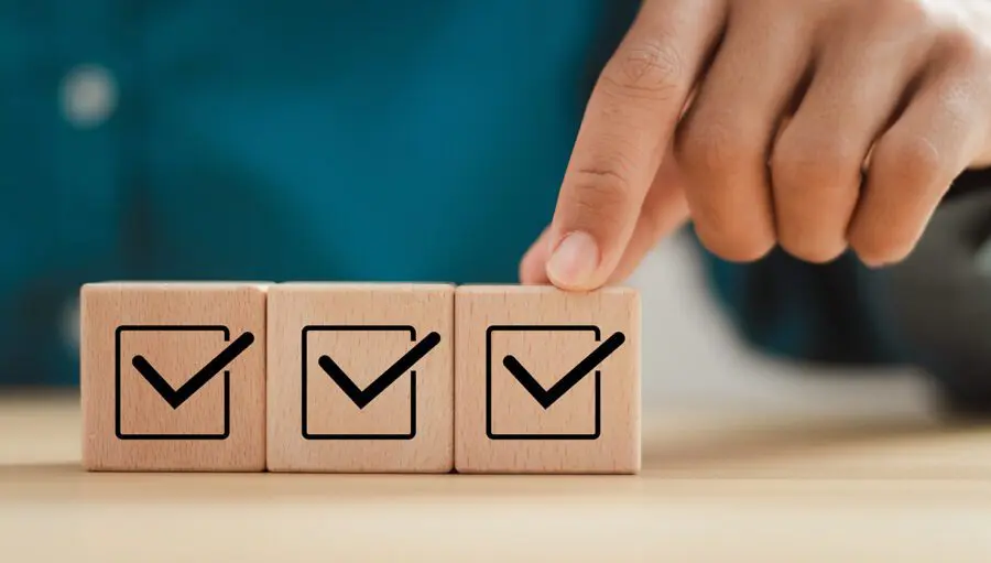 A hand touches a wooden block with a checkmark. Two other blocks with checkmarks are arranged in a row on a table.