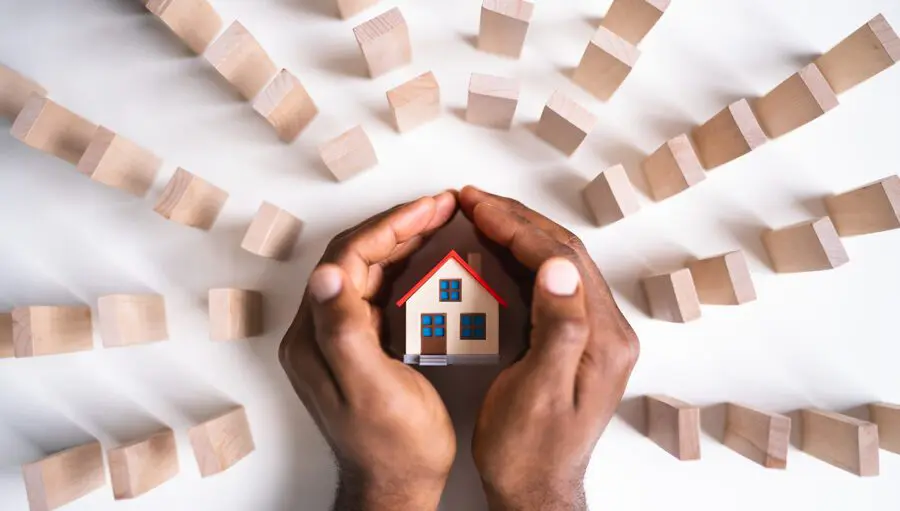 A person’s hands cupping a small model house, surrounded by upright wooden blocks arranged in a semicircle on a white surface.