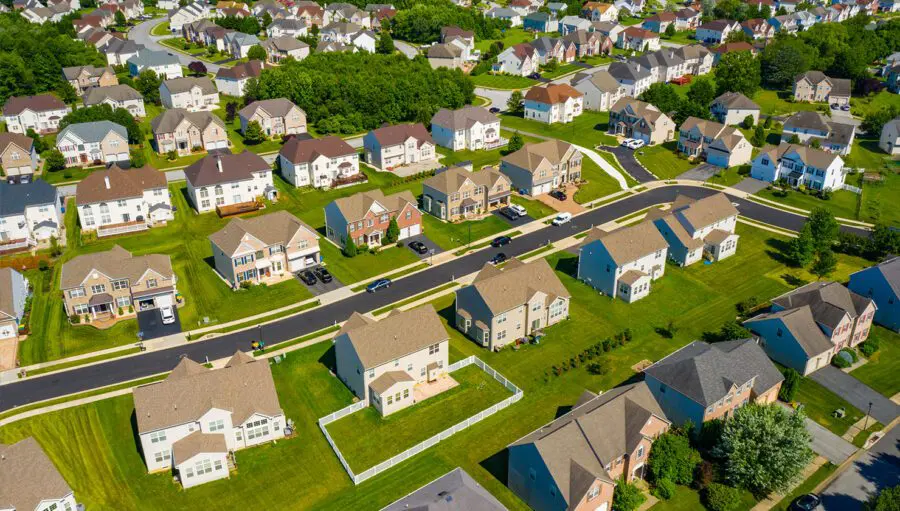 Aerial view of a suburban neighborhood with rows of single-family houses, green lawns, trees, and winding roads on a sunny day.