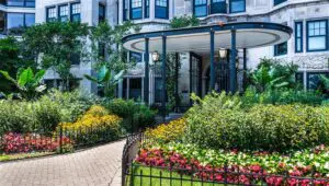 A landscaped garden with colorful flowers surrounds a paved walkway leading to the entrance of a stone building with black columns and decorative details.