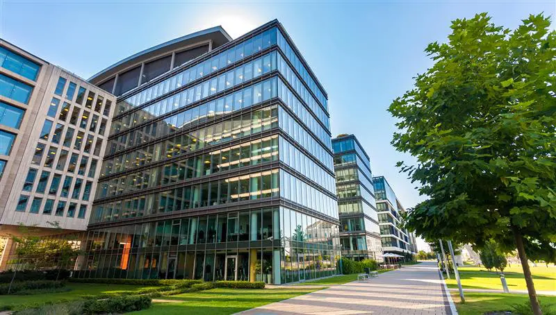 Modern glass office buildings with reflective windows, located alongside a landscaped walkway and green trees on a sunny day.