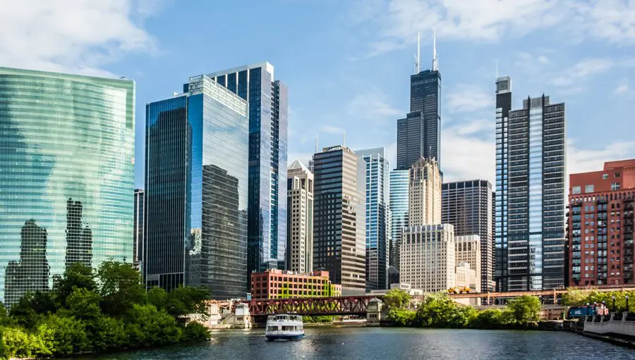 A city skyline with modern glass skyscrapers and older buildings, a river in the foreground, and a boat sailing under a bridge on a partly cloudy day.