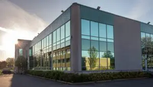 Modern commercial building with large reflective glass windows, concrete walls, and a green roof trim, photographed at sunset with trees and another building visible in the reflection.