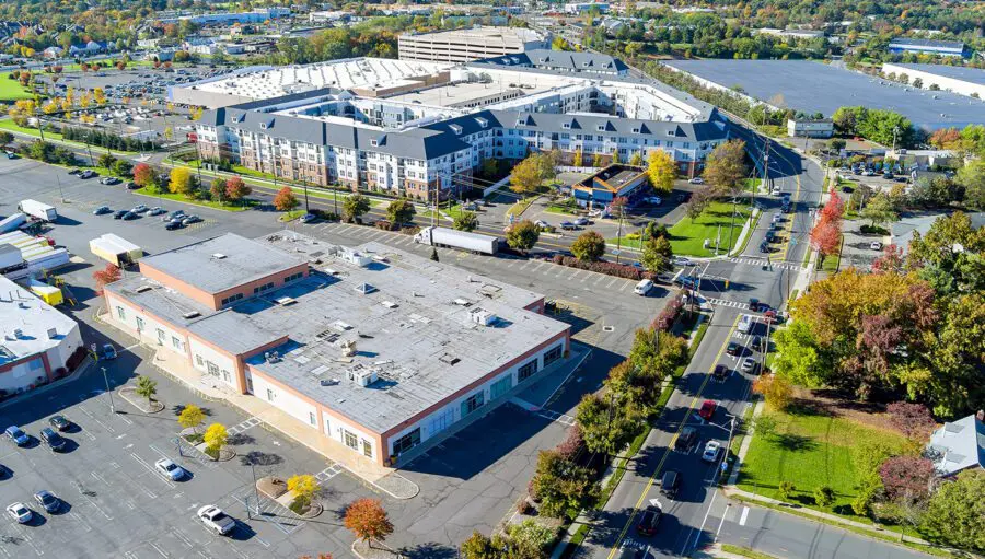 Aerial view of a shopping center parking lot, a large building with a flat roof in the foreground, and apartments with peaked roofs in the background, surrounded by trees and roads.