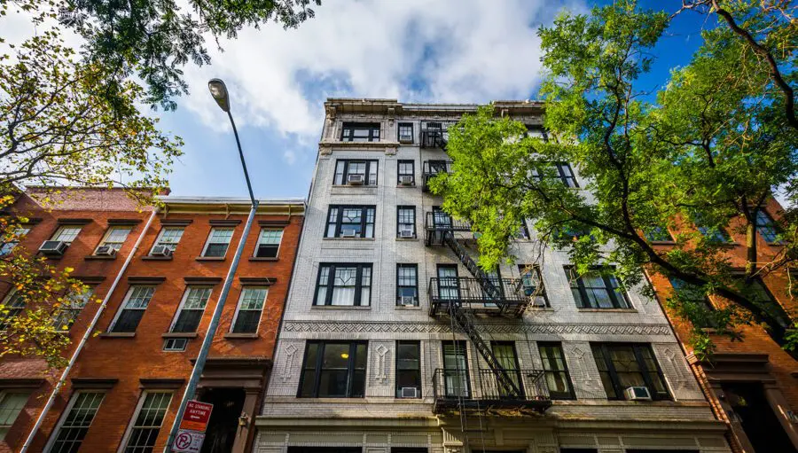 A multi-story brick apartment building with fire escape ladders, framed by trees and a streetlamp, under a partly cloudy blue sky.