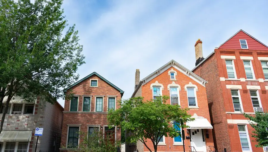 Three brick houses with pitched roofs and various window styles are lined up on a residential street with trees in front and a blue sky overhead.