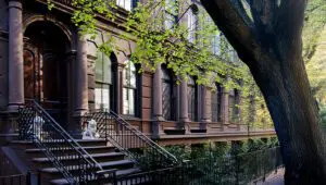 Row of brownstone townhouses with ornate railings, stone steps, and a large tree in the foreground. A white lion statue decorates one of the stoops.