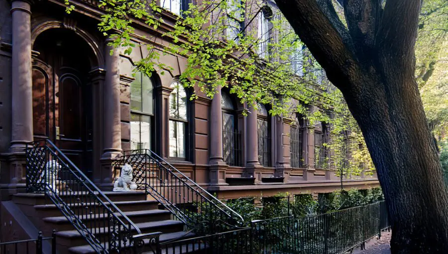 Row of brownstone townhouses with ornate railings, stone steps, and a large tree in the foreground. A white lion statue decorates one of the stoops.
