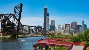 Chicago skyline with Willis Tower in the background, an industrial bridge over the river, kayakers on the water, and a red pedestrian walkway in the foreground.