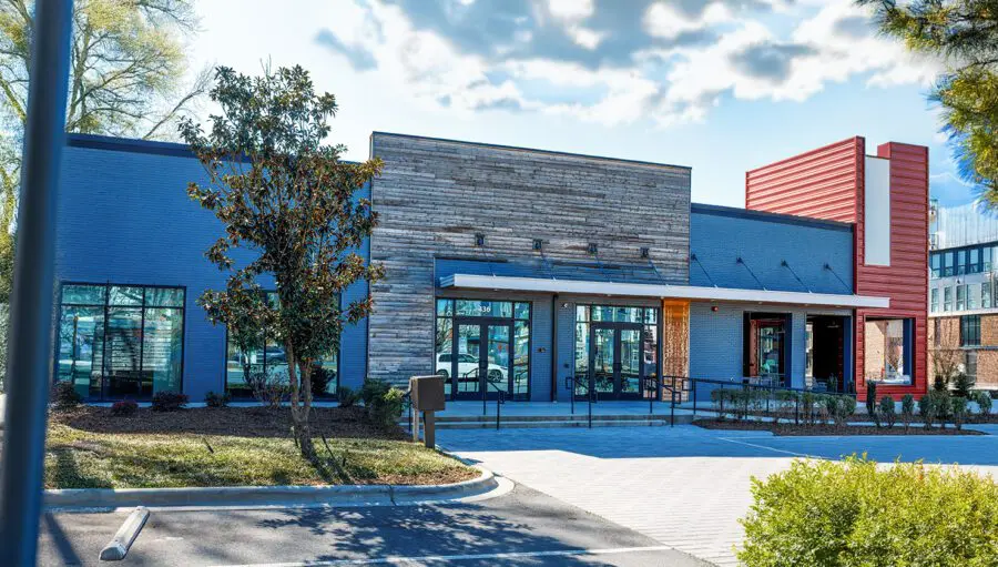 A modern commercial building with large windows, a wooden accent facade, and a red section, set in a landscaped area with a parking lot in front.
