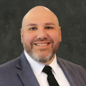 A bald man with a beard is smiling, wearing a gray suit jacket, white shirt, and black tie, in front of a dark textured background.