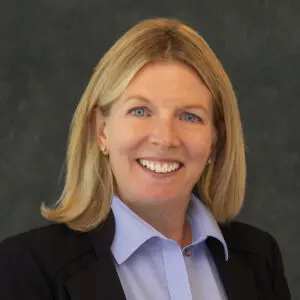 A woman with blonde hair wearing a light blue blouse and dark blazer, smiling, posed in front of a plain dark background.