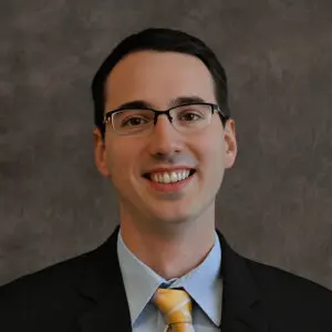 A man wearing glasses, a black suit, light blue shirt, and yellow striped tie, smiling in front of a mottled gray background.
