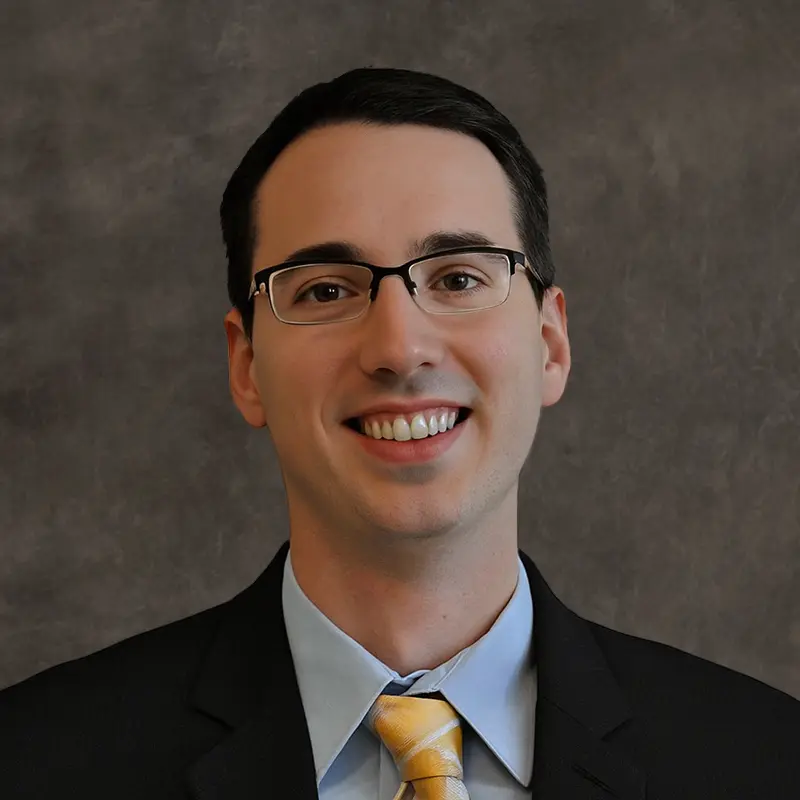 A man wearing glasses, a black suit, light blue shirt, and yellow striped tie, smiling in front of a mottled gray background.