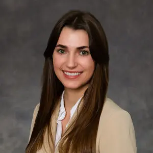 A woman with long brown hair, wearing a beige blazer and white shirt, smiles at the camera in front of a gray background.